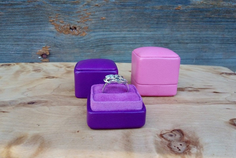May include: A silver ring with a cluster of gemstones sits in a purple velvet-lined ring box. Two additional ring boxes, one purple and one pink, are in the background. The boxes are on a wooden surface.