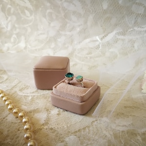 May include: Two rings with green gemstones are displayed in a light pink velvet ring box. The box is sitting on a white lace fabric.