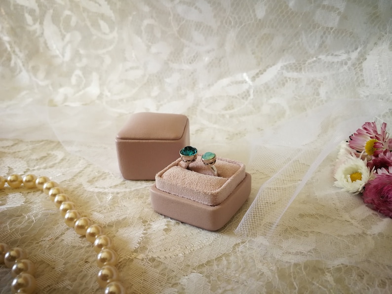 May include: Two rings with green gemstones are displayed in a light pink velvet ring box. The box is sitting on a white lace fabric.