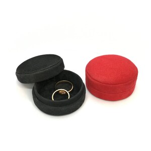 May include: Two gold rings rest inside an open black ring box with a black lid. A red ring box sits to the right. The boxes are round and covered in a textured fabric. The background is white.