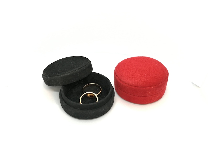 May include: Two gold rings rest inside an open black ring box with a black lid. A red ring box sits to the right. The boxes are round and covered in a textured fabric. The background is white.