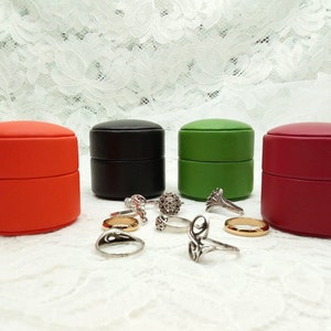 May include: Four small round ring boxes in red, black, green, and burgundy. The boxes are stacked on top of each other. There are several silver rings scattered around the boxes.