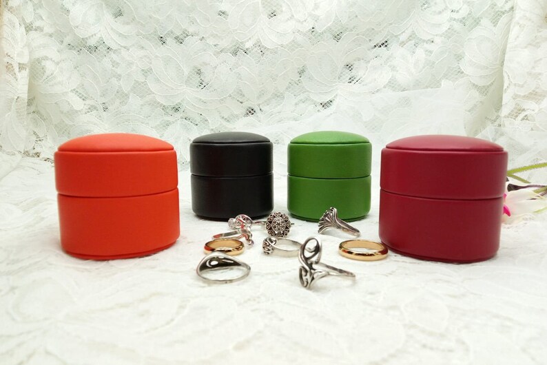 May include: Four small round ring boxes in red, black, green, and burgundy. The boxes are stacked on top of each other. There are several silver rings scattered around the boxes.