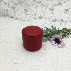 May include: A red, round, two-tiered ring box with a smooth, leather-like finish.
