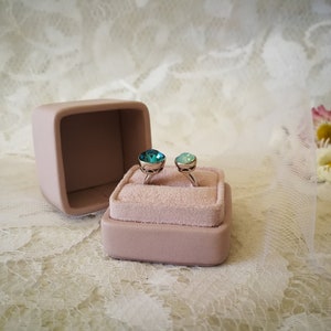 May include: Two rings with blue and green gemstones are displayed in a pink velvet ring box. The box is open and the rings are sitting on a white lace surface.