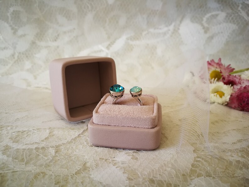 May include: Two rings with blue and green gemstones are displayed in a pink velvet ring box. The box is open and the rings are sitting on a white lace surface.