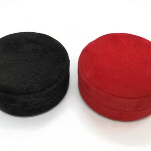 May include: Two round jewelry boxes. One is black, the other is red. Both boxes have a soft, velvety texture and a circular shape. The boxes are closed and appear to be made of a plush material.