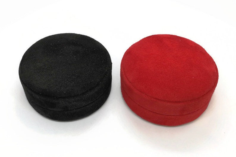 May include: Two round jewelry boxes. One is black, the other is red. Both boxes have a soft, velvety texture and a circular shape. The boxes are closed and appear to be made of a plush material.