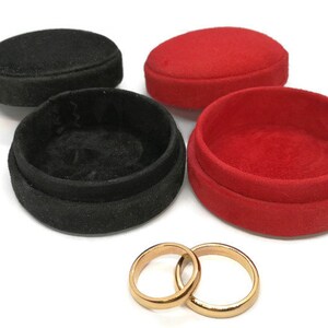 May include: An image of two gold wedding rings next to two open ring boxes, one black and one red. The boxes are round and covered in a soft, velvety material. The rings are simple bands.