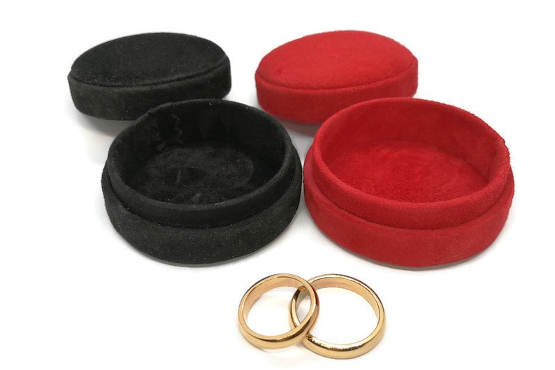 May include: An image of two gold wedding rings next to two open ring boxes, one black and one red. The boxes are round and covered in a soft, velvety material. The rings are simple bands.
