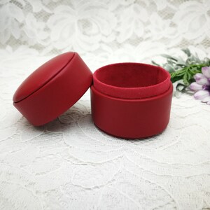 May include: A red leather ring box with a velvet interior. The box is open and the lid is lying next to it.