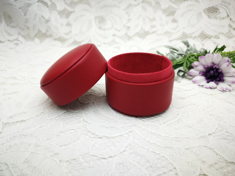May include: A red leather ring box with a velvet interior. The box is open and the lid is lying next to it.