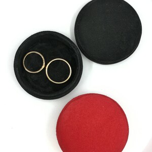 May include: A top-down view of a jewelry tray set. The set includes a black, circular tray with two gold rings inside, a black lid, and a red lid. The items are on a white surface.