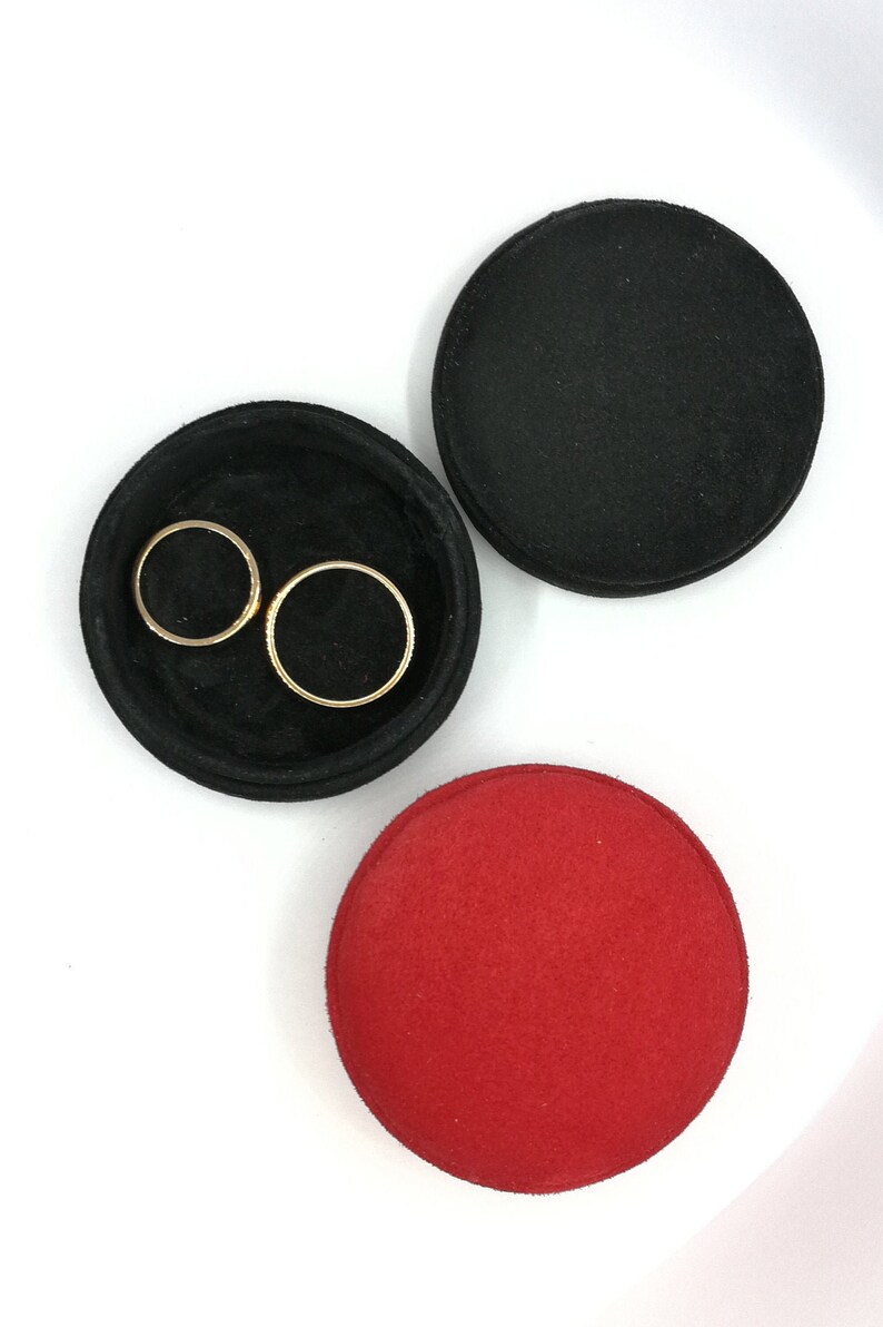 May include: A top-down view of a jewelry tray set. The set includes a black, circular tray with two gold rings inside, a black lid, and a red lid. The items are on a white surface.