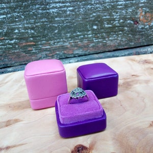 May include: A silver ring with a floral design sits in a purple velvet-lined ring box. Two additional ring boxes, one pink and one purple, are also present. The background features a wooden surface and a weathered wooden panel.