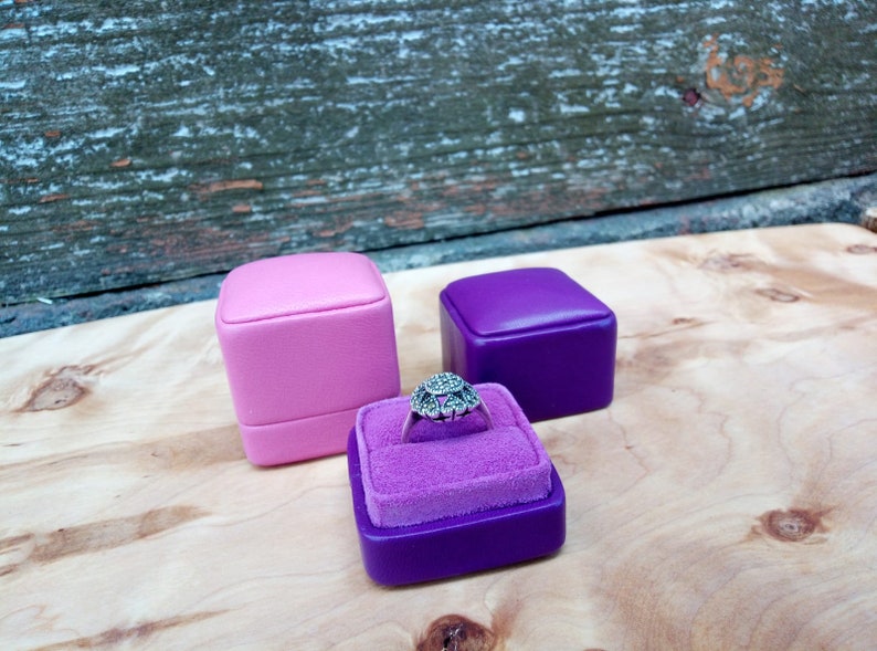 May include: A silver ring with a floral design sits in a purple velvet-lined ring box. Two additional ring boxes, one pink and one purple, are also present. The background features a wooden surface and a weathered wooden panel.