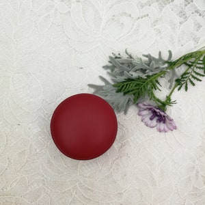 May include: A small, round, red container on a white lace background. A sprig of green and gray foliage with a purple flower is to the right of the container.