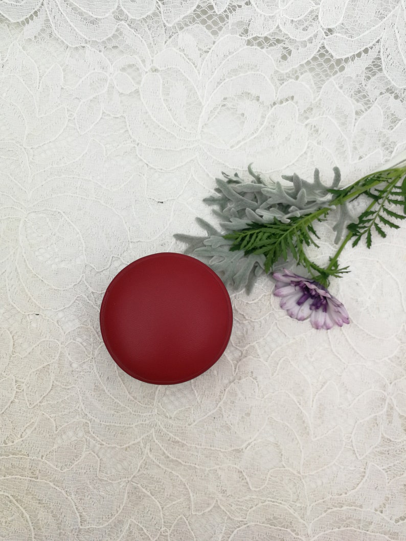 May include: A small, round, red container on a white lace background. A sprig of green and gray foliage with a purple flower is to the right of the container.