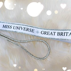 May include: White ribbon keychain with the text "MISS UNIVERSE GREAT BRITAIN" in black. The ribbon is trimmed with silver glitter. A silver rhinestone wrist strap and key rings complete the design.