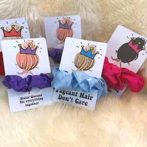 May include: Scrunchies with illustrated cards. The cards feature a cartoon drawing of a bun with a crown. The scrunchies are in purple, light blue, and pink. The cards have text that reads "Sister Queens Tie everything together" and "Pageant Hair Don't Care".