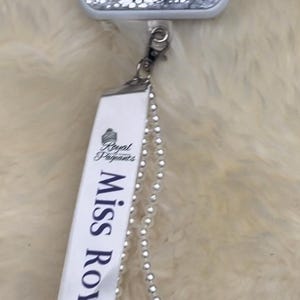 May include: A phone case with a princess design and glitter, attached to a white ribbon with the text "Miss Royal Newcastle" and a pearl necklace.