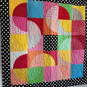 May include: A colorful patchwork quilt with a black and white polka dot border. The quilt features squares and semi-circles in various colors and patterns, including red, pink, yellow, blue, and green. The quilt is hanging on a wooden rod.