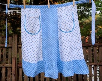 May include: A blue and white floral print apron with a ruffled hem and a large pocket. The apron is hanging on a clothesline.