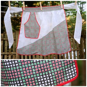 May include: A vintage white apron with a red trim and a pocket. The apron features a checkered pattern in black, red, and green. It is hanging on a clothesline with wooden clothespins. The apron's ties are long and white.