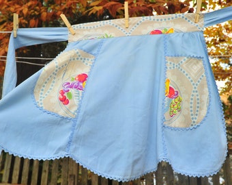 May include: A light blue apron with a scalloped edge and two pockets. The pockets are lined with a cream fabric with a floral print of fruit and leaves.