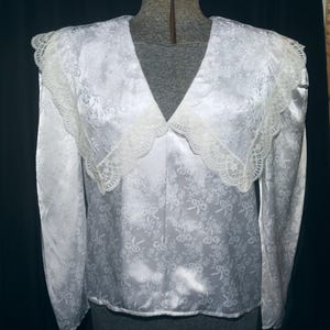 May include: A white long-sleeved blouse featuring a tonal floral and ribbon jacquard pattern, a deep V-neckline, and a wide, scalloped lace collar that extends over the shoulders. The garment has a smooth, satin-like finish and long sleeves.