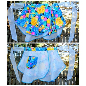 May include: A blue and white floral print apron with a ruffled hem and a single pocket. The apron has two long ties that are tied at the waist.