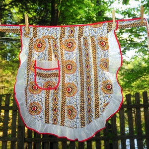 Puede incluir: Un delantal blanco con ribete rojo y un bolsillo blanco. El delantal tiene un estampado africano colorido con formas geométricas amarillas, rojas y marrones. El delantal está colgado en un tendedero.