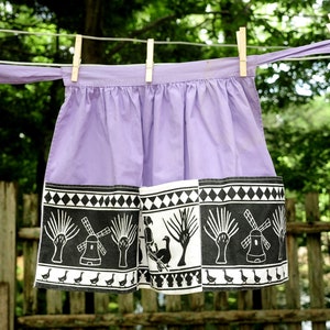 May include: A purple apron with a black and white border featuring a windmill and geese design. The apron has a long tie that hangs down the side.
