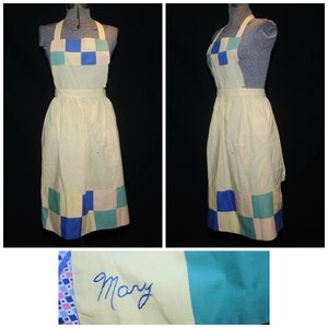 May include: A yellow vintage apron with a blue, green, and pink checkered pattern. The apron has a halter neck strap and a long skirt. The apron is embroidered with the name "Mary".