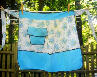 May include: A vintage apron with a white background and blue floral pattern, hanging on a clothesline. The apron features a solid blue top and bottom, a pocket, and black trim. The ties are white with a matching floral pattern.
