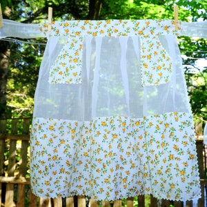 Könnte beinhalten: Eine weiße, durchsichtige Schürze mit gelbem Blumendruck. Die Schürze hat zwei Taschen und einen gewellten Rand.