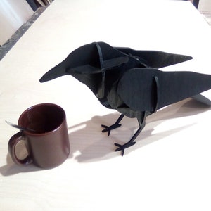 Puede incluir: Una escultura de cuervo de madera negra con alas y patas detalladas. El cuervo está posado sobre una superficie de madera clara junto a una taza de cerámica marrón.