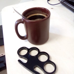 May include: A brown ceramic mug filled with coffee and a black metal knuckle duster on a white surface.