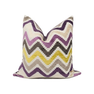 May include: A decorative throw pillow with a chevron pattern. The pillow features a repeating zigzag design in shades of purple, yellow, gray, and beige against an off-white background. The pillow is square and has a soft, textured appearance.