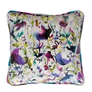 May include: Square decorative pillow with a floral pattern in shades of purple, blue, green, and yellow on a light gray background. The pillow has a contrasting trim in shades of purple and blue. The design features watercolor-style flowers and foliage.