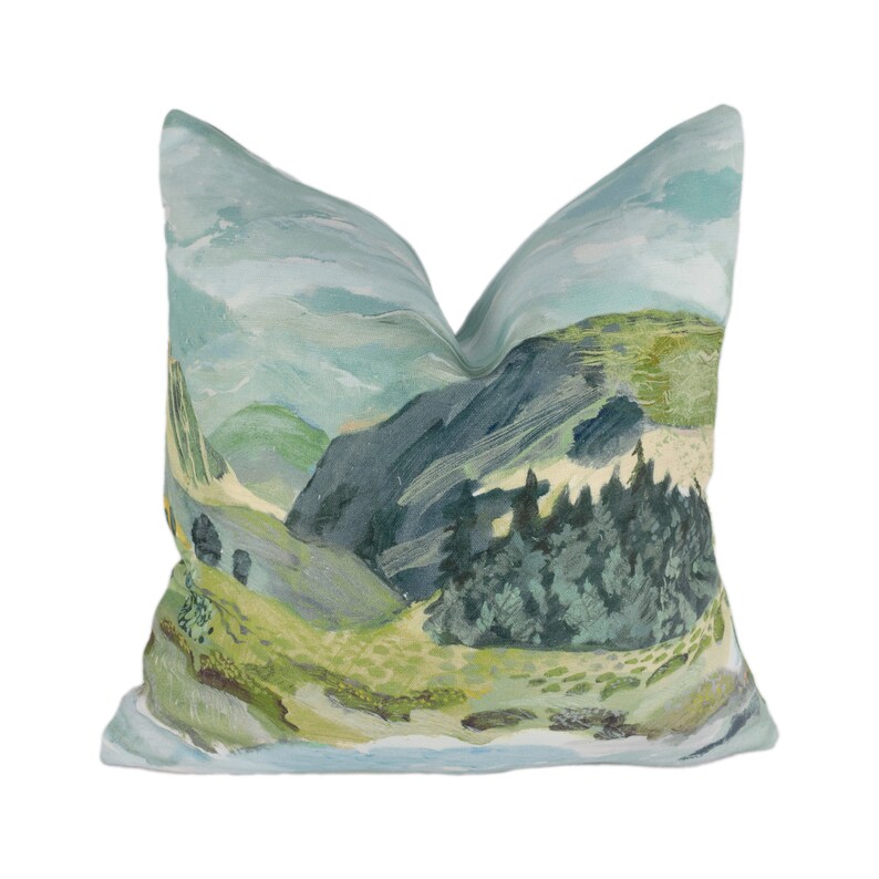 May include: A decorative cushion featuring a landscape design with a mountain range, trees, and a sky with clouds. The cushion is made from a soft, light blue fabric.