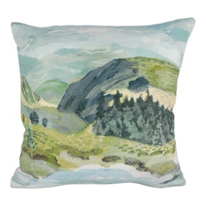 May include: A decorative throw pillow featuring a watercolour-style painting of a mountain landscape. The pillow has a blue, green and white colour scheme with a depiction of a mountain range, trees and a cloudy sky.