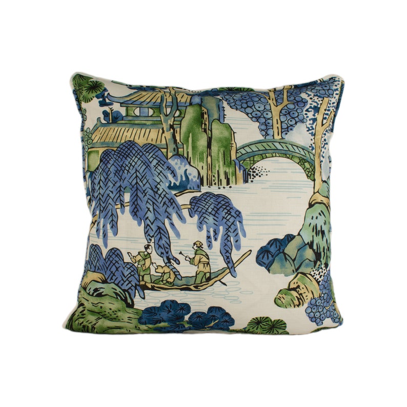 May include: A decorative throw pillow with a blue and green landscape design featuring a bridge, trees, and figures in a boat. The background is a light cream color.