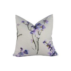 Op de afbeelding: Een vierkant decoratief kussen met een bloemenontwerp. Het kussen heeft een lichtbeige achtergrond met een aquarel-achtige print van paarse bloemen en donkergrijze stelen. Een zacht, decoratief item.
