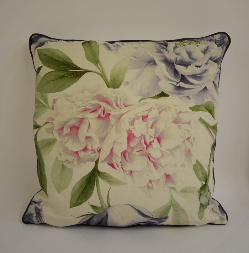 Puede incluir: Coj&iacute;n decorativo cuadrado con un gran estampado floral de peon&iacute;as en tonos blancos, rosas y morados. El coj&iacute;n tiene un borde azul marino oscuro y hojas verdes. Forma cuadrada.