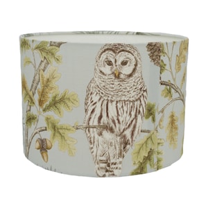 May include: A light blue lampshade with a brown and white owl perched on a branch with green leaves. The owl has a detailed design and is the focal point of the lampshade.