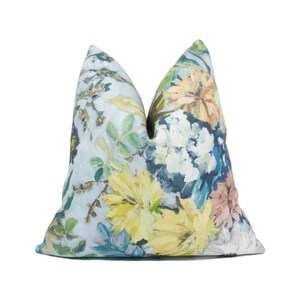 May include: A square decorative cushion with a floral design. The cushion has a light blue backdrop with a painted pattern of yellow, blue, and peach flowers and green leaves. The cushion is a square shape.