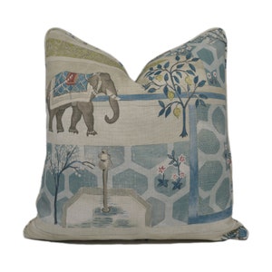 Puede incluir: Una almohada decorativa con un fondo azul claro y beige que presenta una ilustración estilizada de un elefante, un limonero y una fuente. El diseño recuerda al arte asiático tradicional.