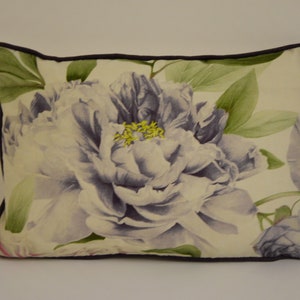 Puede incluir: Coj&iacute;n decorativo rectangular con una gran peon&iacute;a de estilo acuarela en tonos lavanda y blanco. El dise&ntilde;o floral se acent&uacute;a con hojas verdes y estambres amarillos, sobre un fondo crema. Tiene un borde morado oscuro.