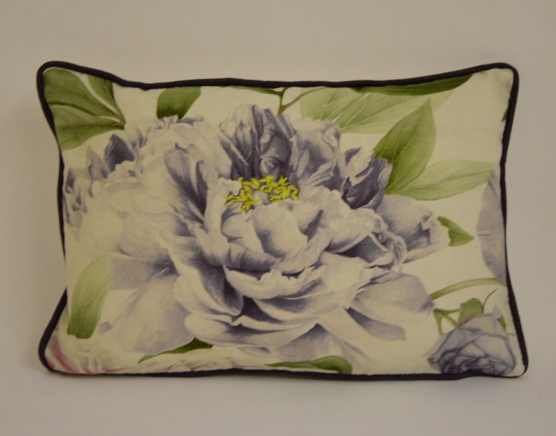 Puede incluir: Coj&iacute;n decorativo rectangular con una gran peon&iacute;a de estilo acuarela en tonos lavanda y blanco. El dise&ntilde;o floral se acent&uacute;a con hojas verdes y estambres amarillos, sobre un fondo crema. Tiene un borde morado oscuro.